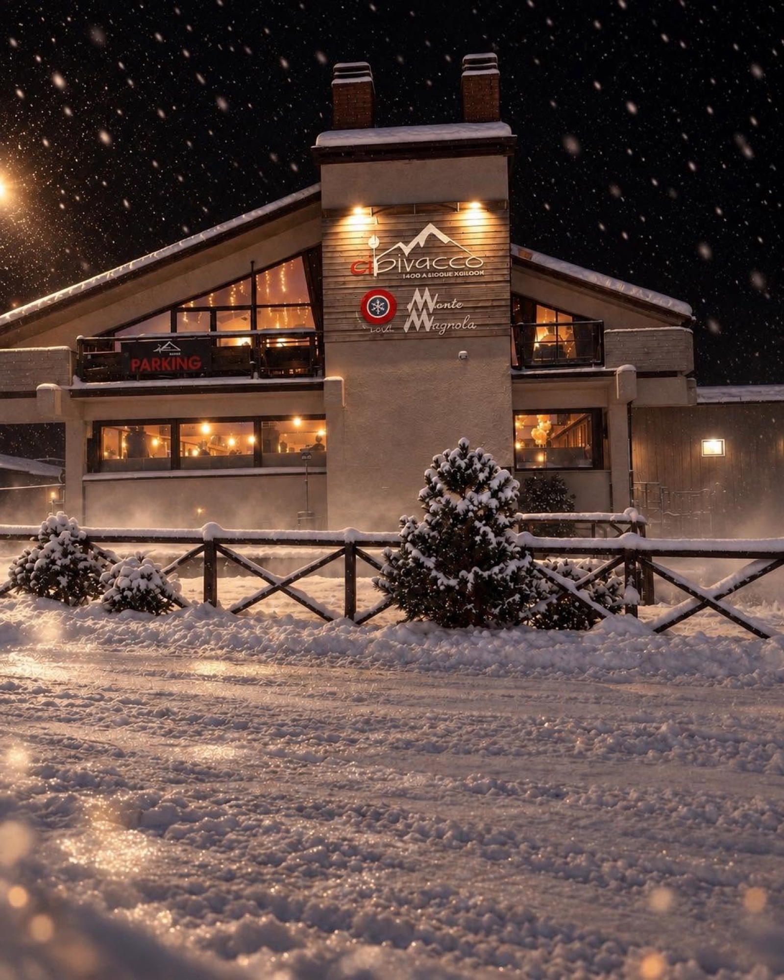 La facciata del Cibivacco di notte con la neve che cade, il parcheggio e le finestre illuminate